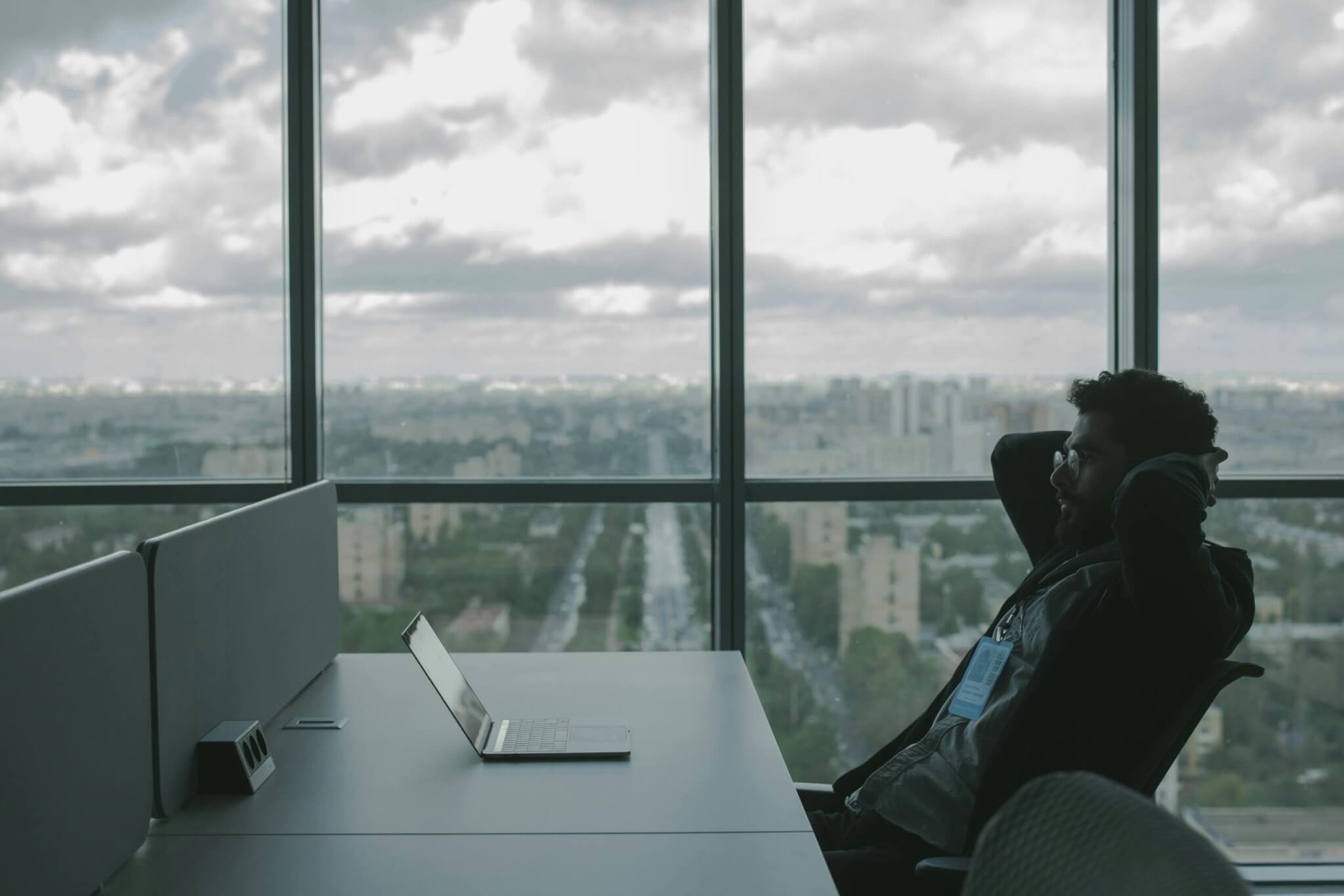A man sits at his desk in front of his laptop. He has his hands crossed behind his head as if in frustration.