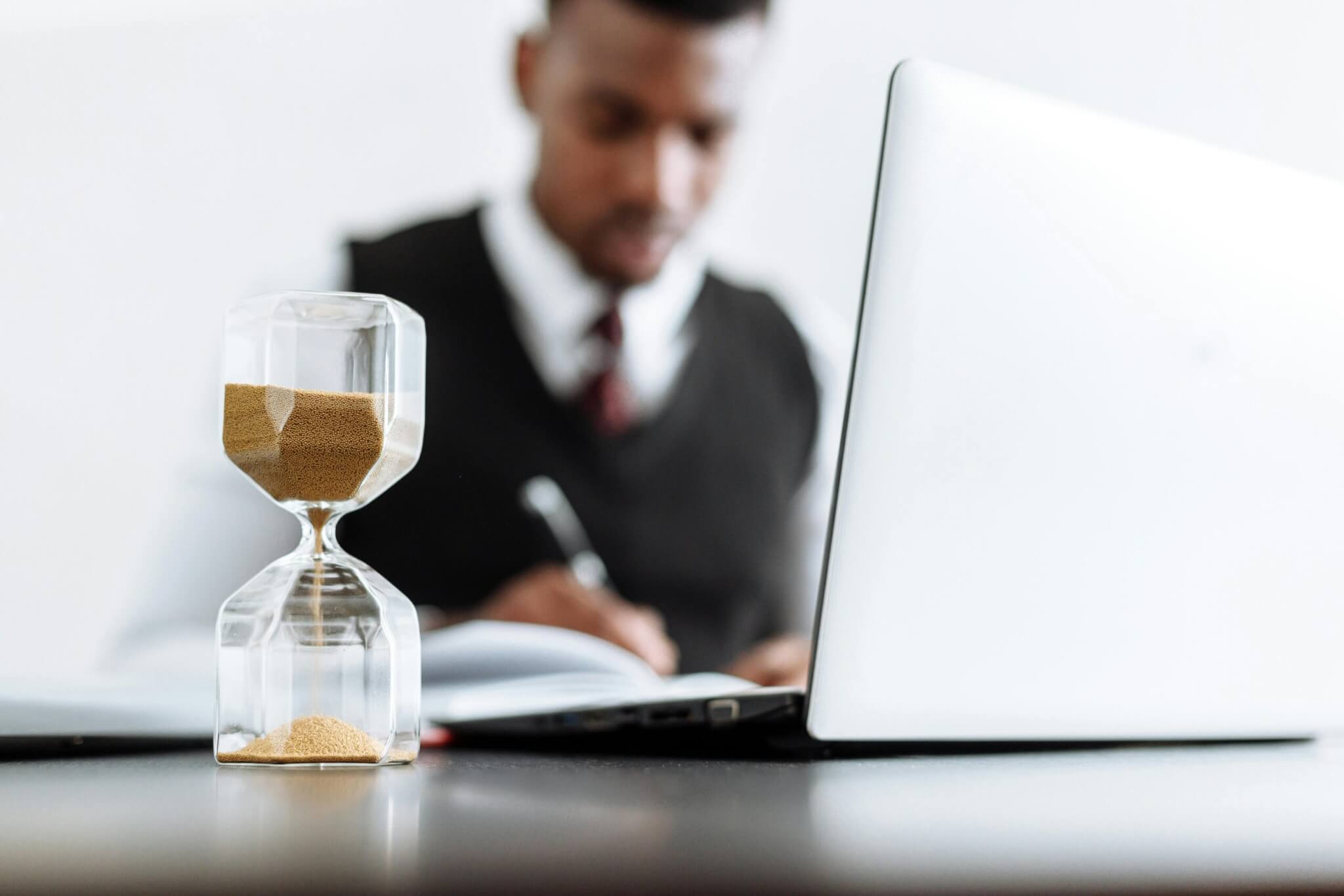 A man sits at a desk with his laptop and a notepad. A sand timer is in focus in the foreground.