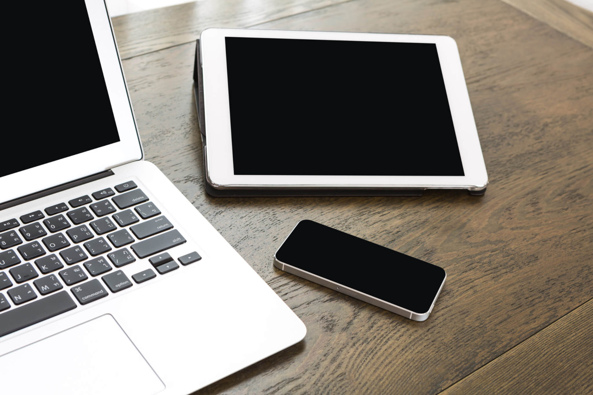 A wooden table with an iPad, iPhone and partially visible Macbook. Their screens are blank.
