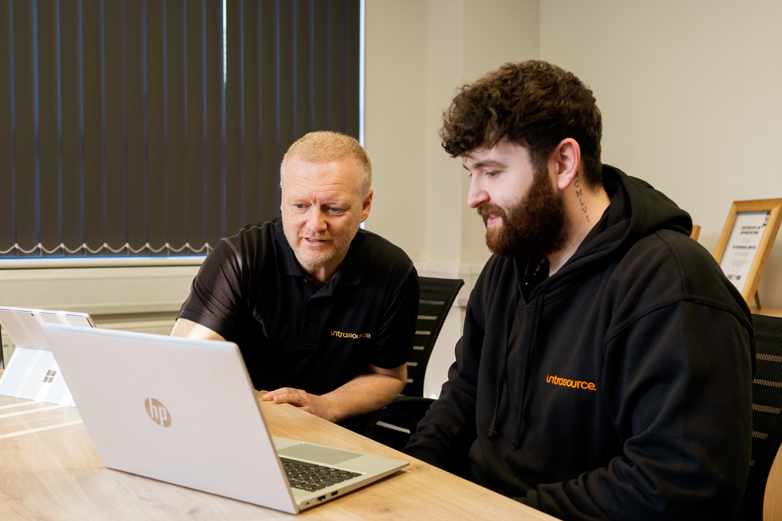 Two men in black Intrasource uniforms look towards a silver laptop. They're mid discussion.