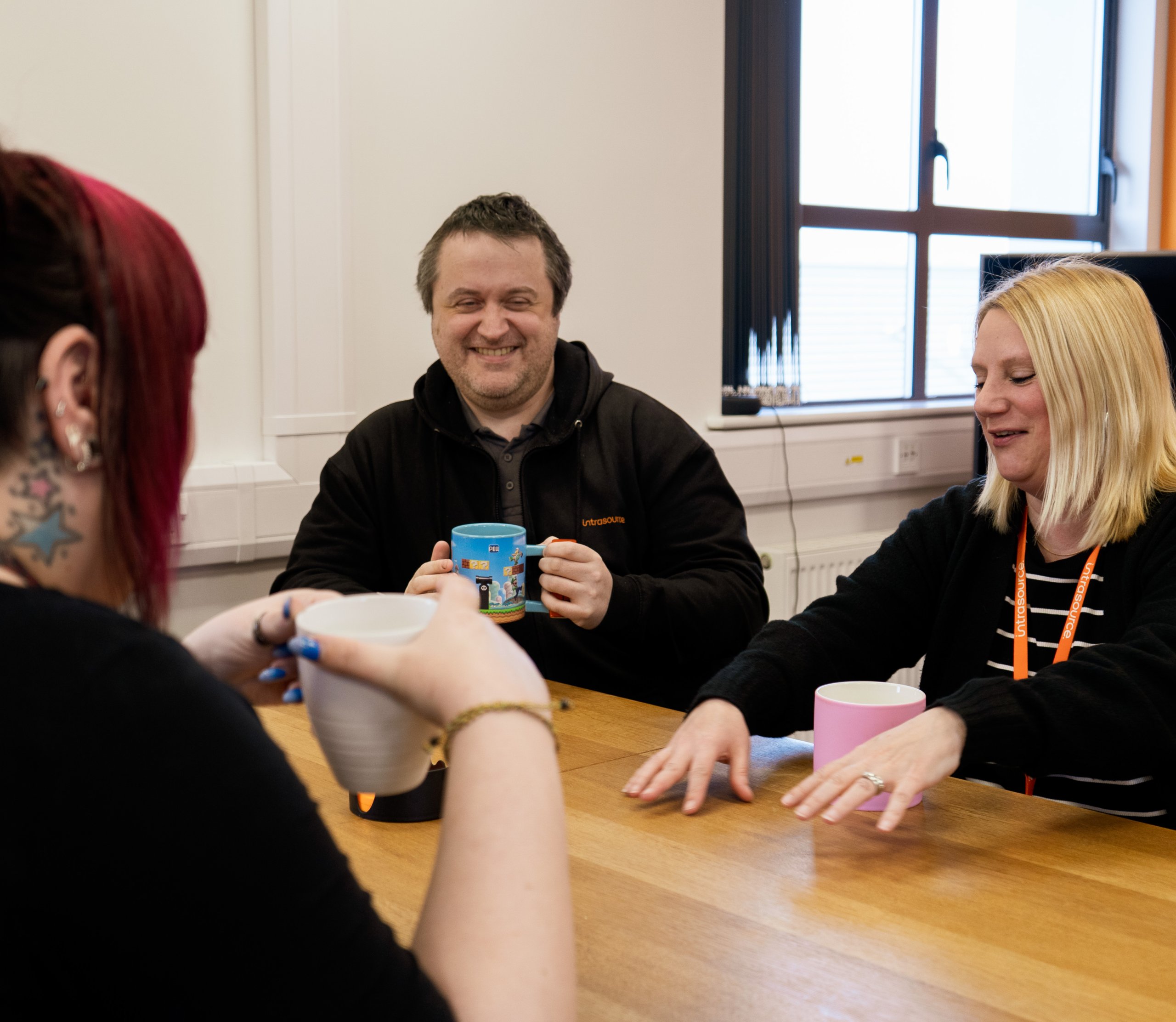 3 Office workers sit around an office desk laughing and holding mugs.